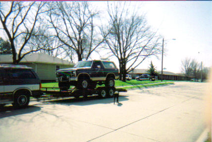 1986 Ford Bronco - A dark-colored Ford Bronco is loaded onto a trailer, towed by a silver SUV on a suburban street.