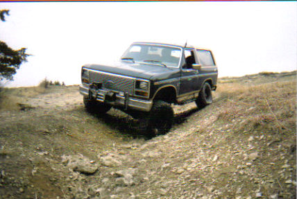 1986 Ford Bronco - A dark blue 1980 Ford Bronco navigates an off-road trail on a dry, grassy hill under a bright sky.