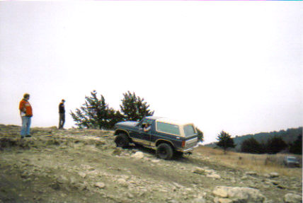 1986 Ford Bronco - A dark blue SUV with a white top navigates a rocky, off-road path with two people observing.
