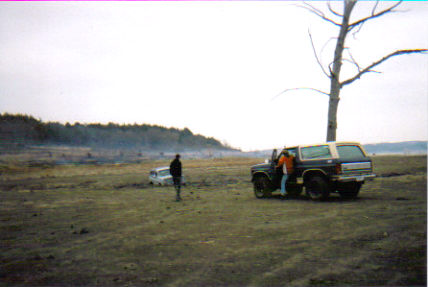 1986 Ford Bronco - A Ford Bronco is parked in a field with a person entering the driver's side, while another vehicle and person are visible in the background.