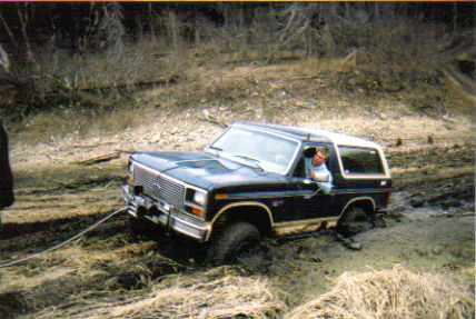 1986 Ford Bronco - A 1986 Ford Bronco is seen stuck in muddy terrain, with a tow rope attached and a driver in the cab.