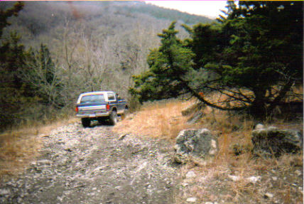 1986 Ford Bronco - A blue and white 1986 Ford Bronco navigates a rough, unpaved path through a wooded, hilly area.