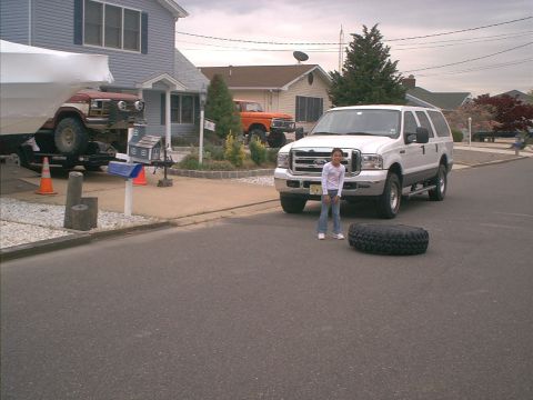 1978 Ford Bronco - A white SUV is parked in a driveway next to a girl and a spare tire, with a red Jeep on a trailer behind it.