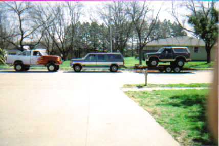 1986 Ford Bronco - A lifted Ford Bronco is transported on a trailer, accompanied by a pickup truck and a large SUV.