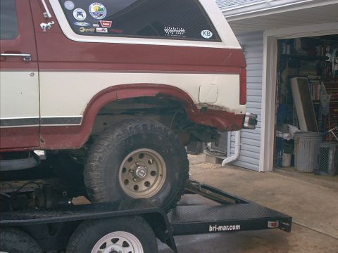 1986 Ford Bronco - A red and white SUV is loaded onto a trailer, with a garage visible in the background.