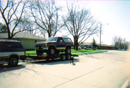 1986 Ford Bronco - A dark 1986 Ford Bronco is transported on a trailer, with another vehicle visible on the left.