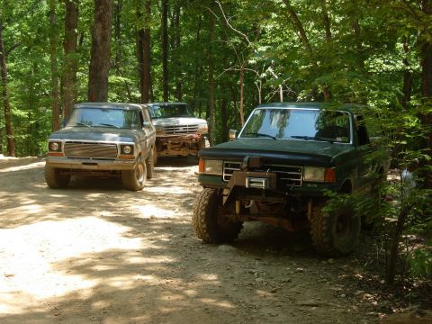 1995 Ford Bronco - Three off-road vehicles navigate a wooded trail, with a dark green Bronco leading the pack.