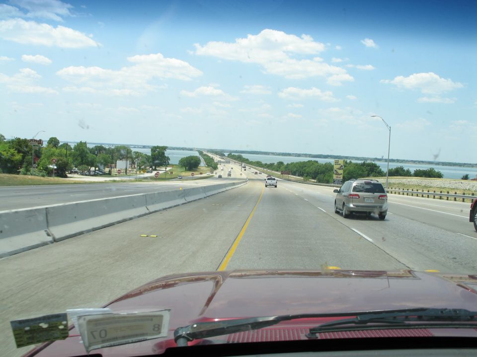 Red vehicle's dashboard and windshield view of a highway with a silver SUV and other cars ahead, under a blue sky with clouds