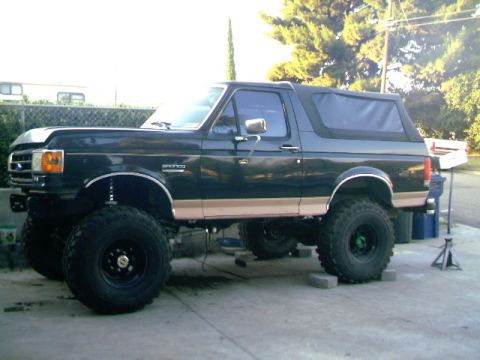 1989 Ford Bronco - This lifted black Ford Bronco features a tan side trim and is shown from a 3/4 front angle.