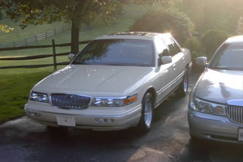 1996 Mercury Grand Marquis - A 1996 Mercury Grand Marquis is parked on a wet driveway with a wooden fence and trees in the background.