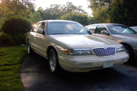 1996 Mercury Grand Marquis - A tan sedan is parked next to another car on a sunny day, with trees and bushes in the background.