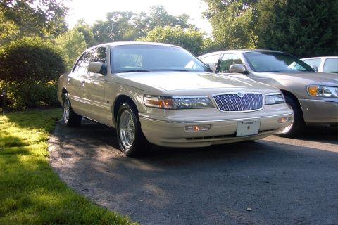 1996 Mercury Grand Marquis - A tan sedan is parked on an asphalt driveway with trees and grass in the background.