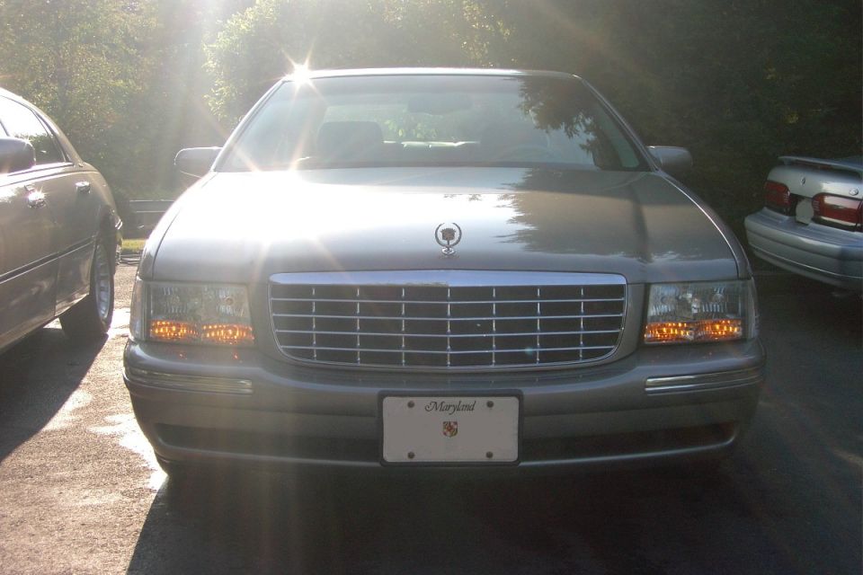 Silver Cadillac sedan front view with chrome grille and Maryland plate, backlit by sun.