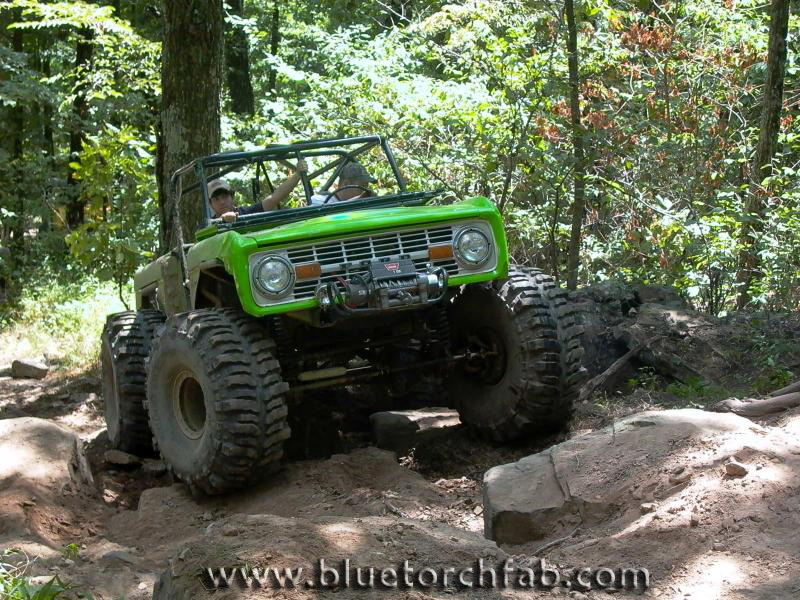 Lime green early Ford Bronco on huge mud tires crawling a rock ledge on a wooded trail