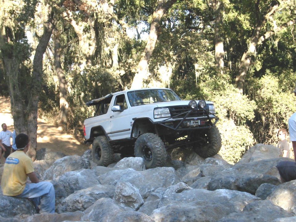 White full-size Ford Bronco with winch bumper rock-crawling over granite boulders as spectators watch