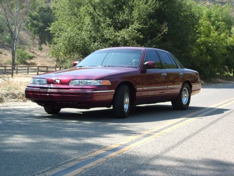 1992 Ford Crown Victoria - A maroon 1992 Ford Crown Victoria sedan is parked on the side of a road with trees in the background.