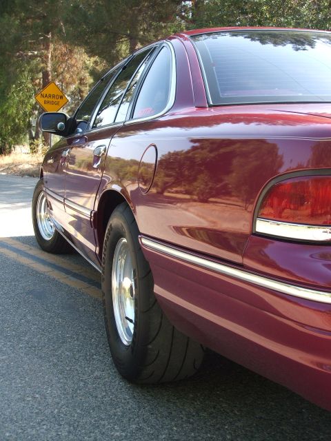 1992 Ford Crown Victoria - A maroon sedan with wide rear tires is parked on the side of a road near a narrow bridge sign.