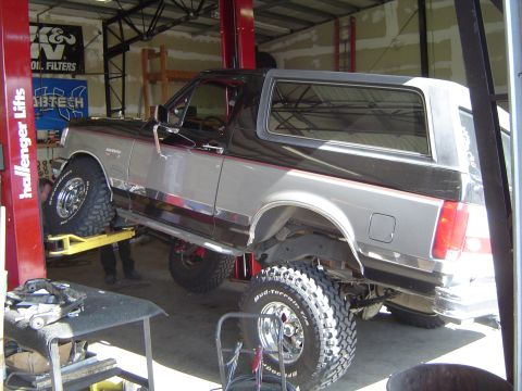 1989 Ford Bronco - A two-tone Ford Bronco is raised on a hydraulic lift in a workshop, with its rear suspension and large tires visible.