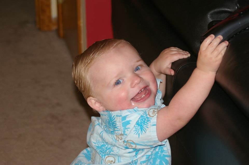 Baby with blonde hair and blue eyes wearing a blue patterned shirt reaches for a black object