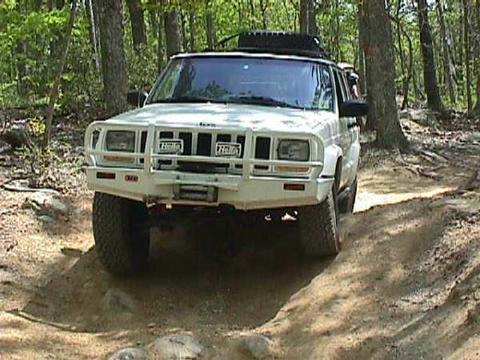 1996 Ford Bronco - A white Jeep Cherokee XJ navigates a rocky, wooded trail with a brush guard and roof rack.