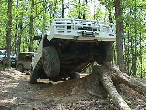 1996 Ford Bronco - A white off-road truck navigates a challenging obstacle, climbing over a large fallen tree in a wooded area. Another vehicle is visible in…