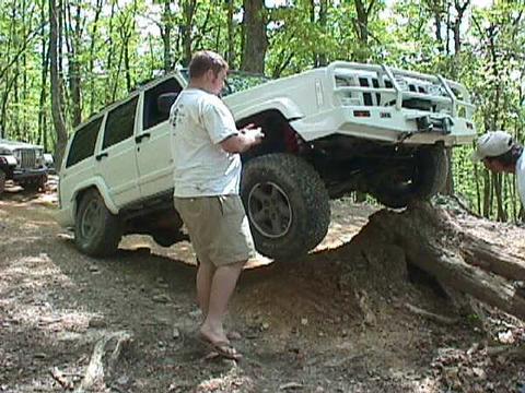 1996 Ford Bronco - A white Jeep Cherokee XJ navigates an obstacle on a wooded trail, with another vehicle in the background.