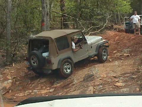1996 Ford Bronco - A tan Jeep Wrangler navigates a wooded dirt trail with another vehicle visible in the background.