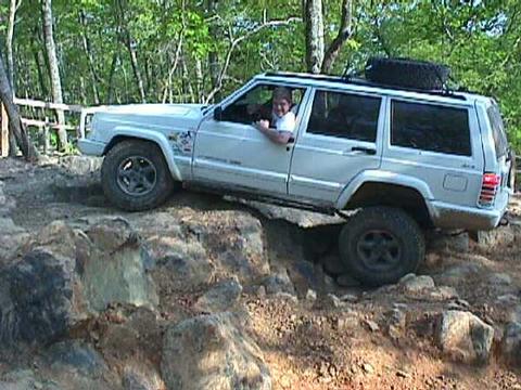 1996 Ford Bronco - A white Jeep Cherokee XJ navigates a rocky off-road trail with a spare tire mounted on its roof.