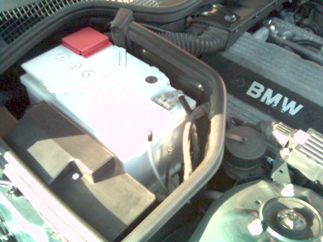 Engine bay with white battery and red terminal cover next to BMW-labeled valve cover