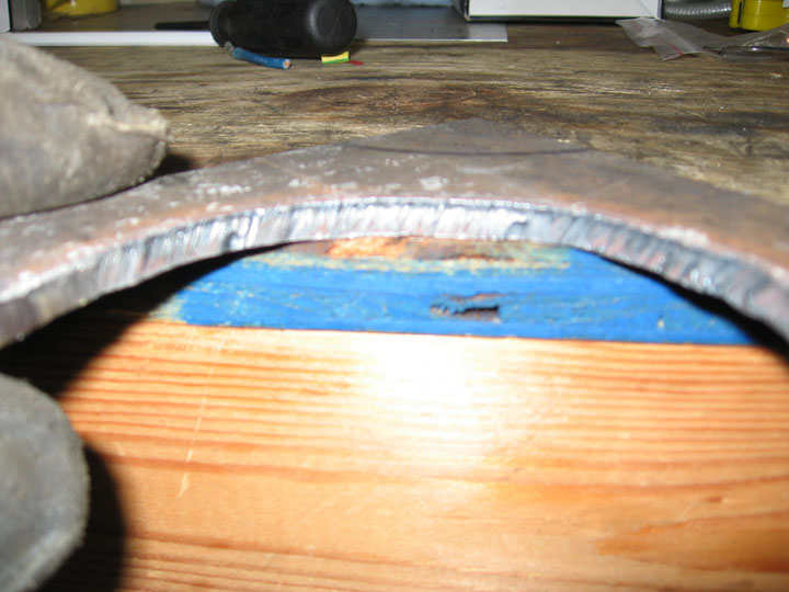 Close-up of a curved metal piece with rough edges, resting on a wooden surface with a blue shim underneath