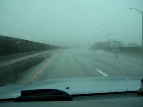 1992 Ford Taurus - Driver's-eye view through a rain-streaked windshield on a multi-lane highway in heavy rain, with the car's light-colored hood and dashboard…
