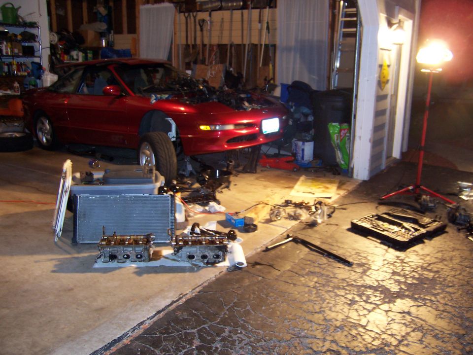 Red coupe with hood open, engine parts and radiator on the floor in a garage