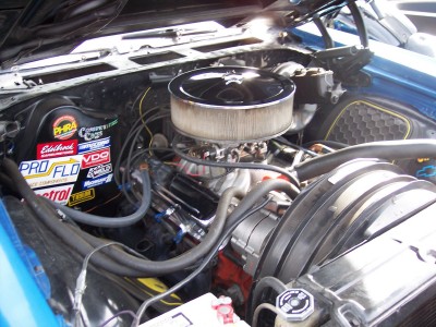 1972 Ford Ranchero - The engine bay of a blue car is shown, various performance parts and stickers on the fender well.