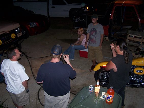 People gathered around vehicles and a motorcycle at night, with drinks on a table