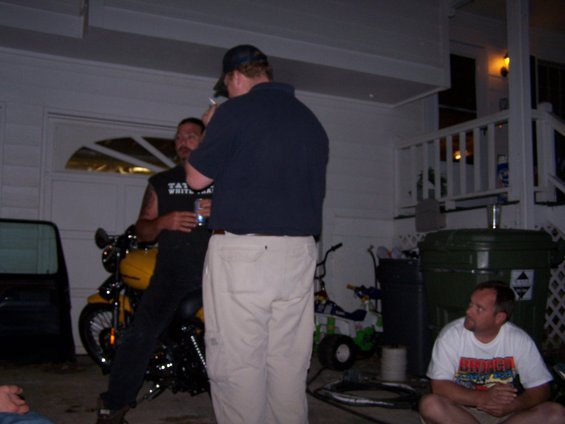 Guy in black shirt leans on a yellow motorcycle, talking to a guy in a blue shirt. Another guy sits on the ground nearby