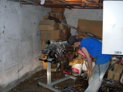 1992 Ford Taurus - A V6 engine mounted on a gray engine stand in a cluttered basement shop, with rusty exhaust manifolds and intake plumbing still attached. A…