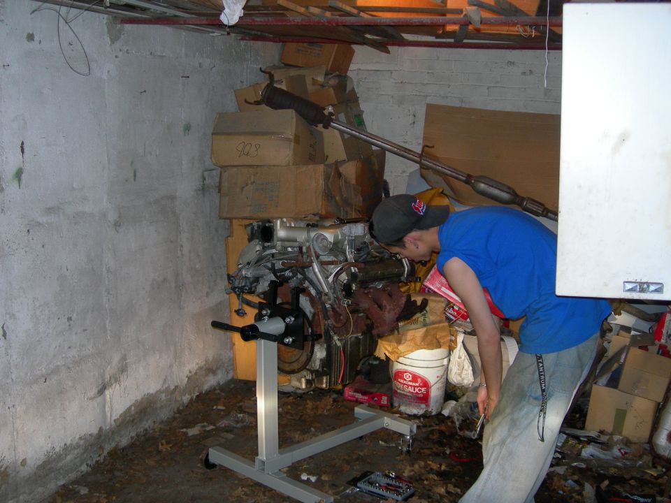 V6 engine on engine stand in basement shop while a person works on it with a wrench