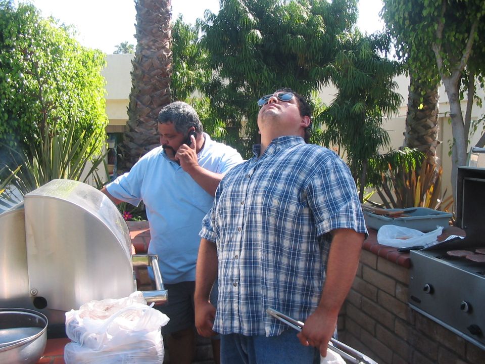 Two men stand by barbecues outdoors, one on a phone, the other looking up