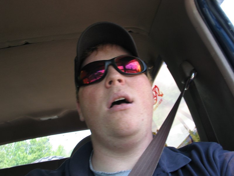 Person in cap and sunglasses seated in a car with tan headliner and seat belt