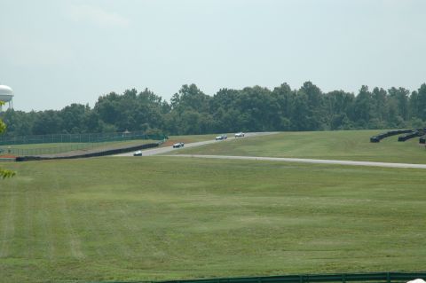 2000 Ford Mustang - Cars are seen on a race track during a sunny day with a treeline and water tower visible.