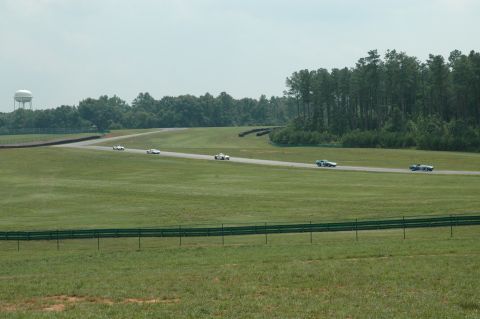 2000 Ford Mustang - Multiple vehicles are seen racing on a track with a water tower and trees in the distance.