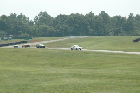 2000 Ford Mustang - Multiple vintage race cars compete on a sunny day at a racetrack, with trees and grassy hills in the background.