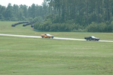 2000 Ford Mustang - Two classic muscle cars compete on a paved racetrack surrounded by grassy fields and trees.