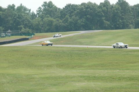 2000 Ford Mustang - Three vintage Mustangs race on a sunny day at a track, with a yellow Mustang in the foreground.