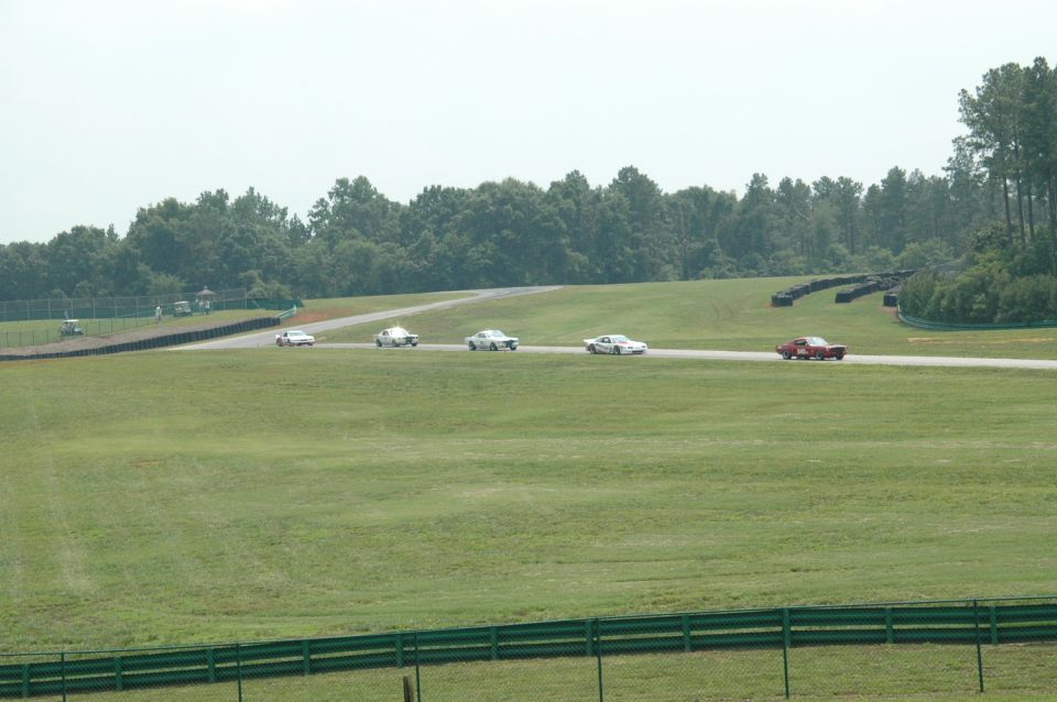 Several vintage cars race on a track with green grass and trees in the background
