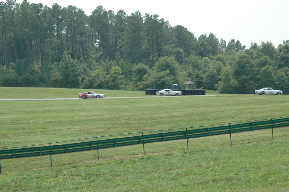 Three cars racing on a track with trees and a fence in the foreground