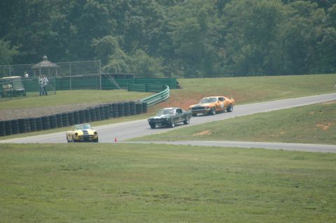 2000 Ford Mustang - A yellow, black, and orange race car compete on a track with a wooded background.