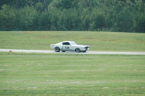 2000 Ford Mustang - A classic white Mustang with racing numbers drives on a paved track surrounded by green grass and trees.