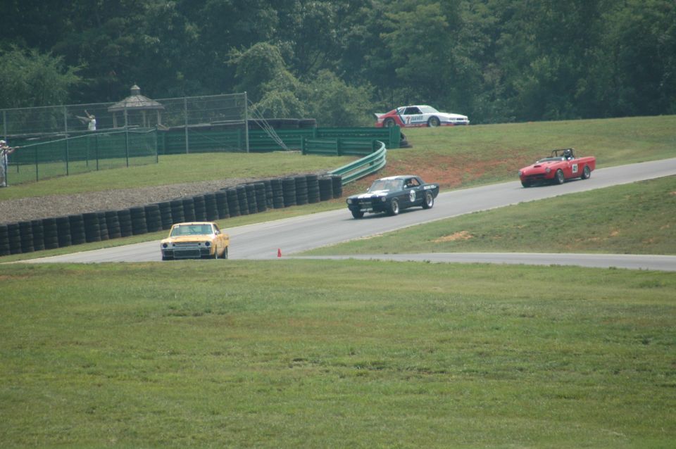 Several cars race on a track with grassy hills and trees in the background