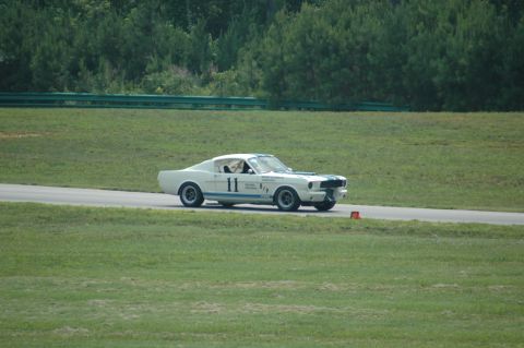 2000 Ford Mustang - A classic Mustang Shelby GT350-style race car is seen on a track, identified by its distinctive body style and racing decals.